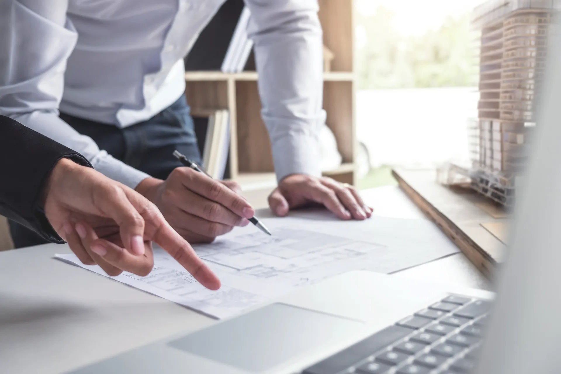 Hände zeigen auf technische Zeichnungen auf einem Schreibtisch neben einem Laptop.